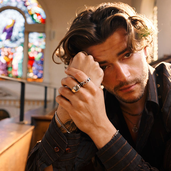 Man with wavy hair rests head on hands with rings sitting in a room with stained glass windows