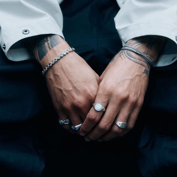 Male hands holding each other wearing 4 Silver rings and multiple Silver bracelets on a dark background