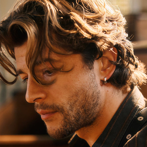 Close-up of a man with wavy hair and a black shirt, with a blurred background with a Silver hoop in his ear lobe.