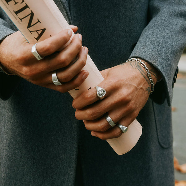 Model wearing a grey coat holds a newspaper with both hands, wearing multiple Silver rings and a Silver bracelet