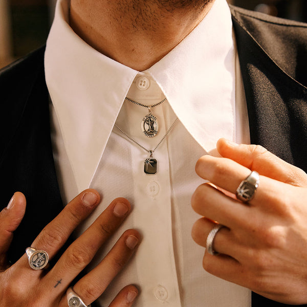 Model wearing a white shirt with 2 pendant necklaces under the collar and silver rings on his hands