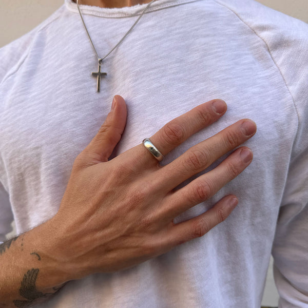 Model wears a white top with a Silver cross necklace over it and on his index finger is a silver band ring with a green gemstone - Serge DeNimes